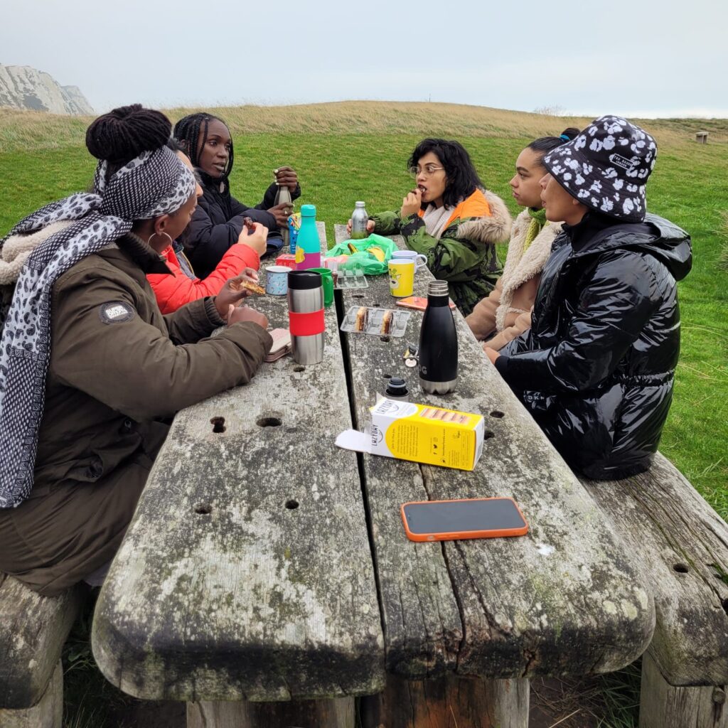 A group of six women sat around a wooden table having a conversation. In the background you can see grass, cliffs and blue sky.