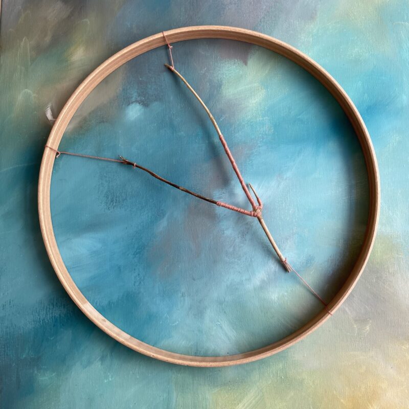 A thin twig wrapped in coral pink thread set inside a wooden hoop. The background is an abstract painting in tones of blue and brown.