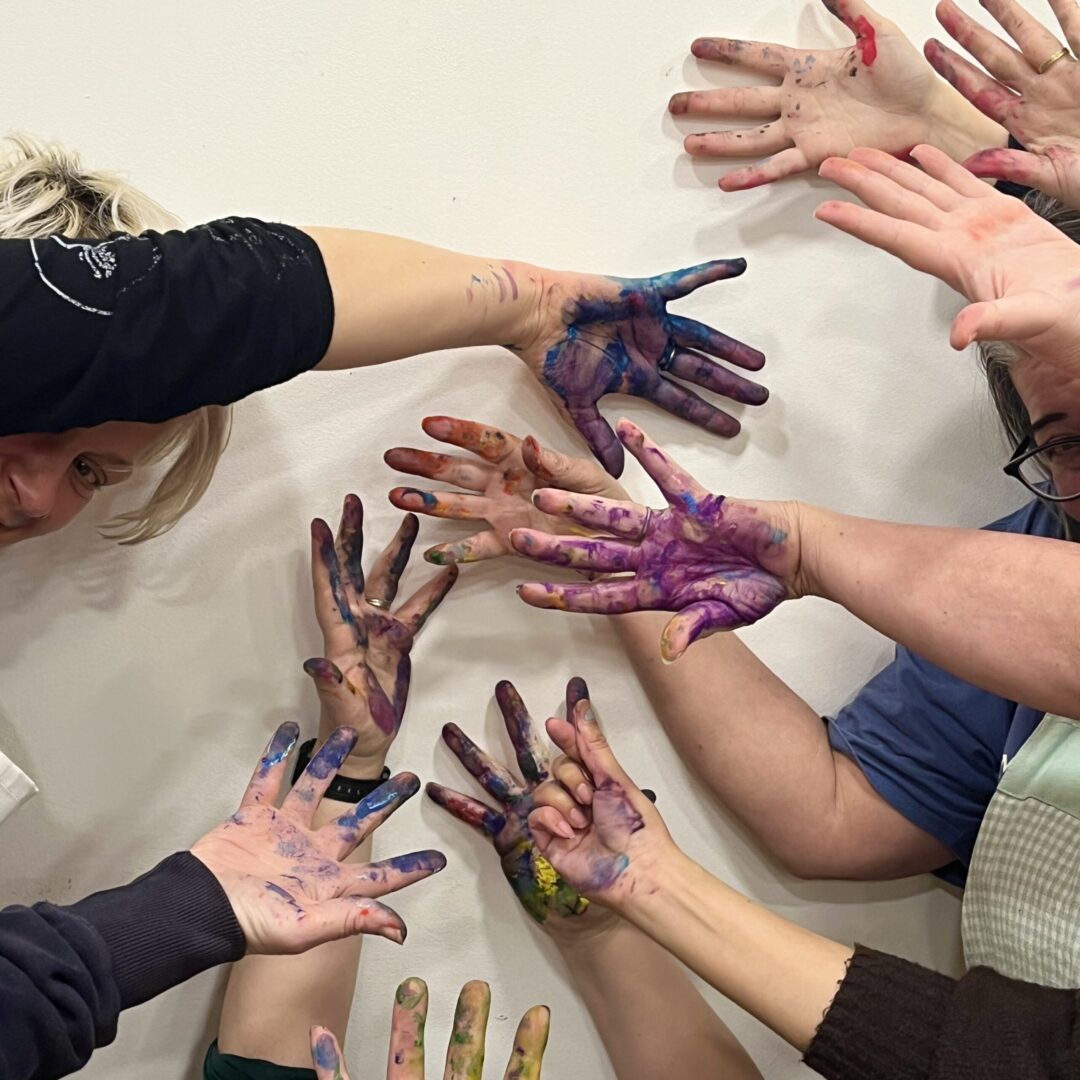 A group of people holding their brightly coloured paint covered hands against a white wall.
